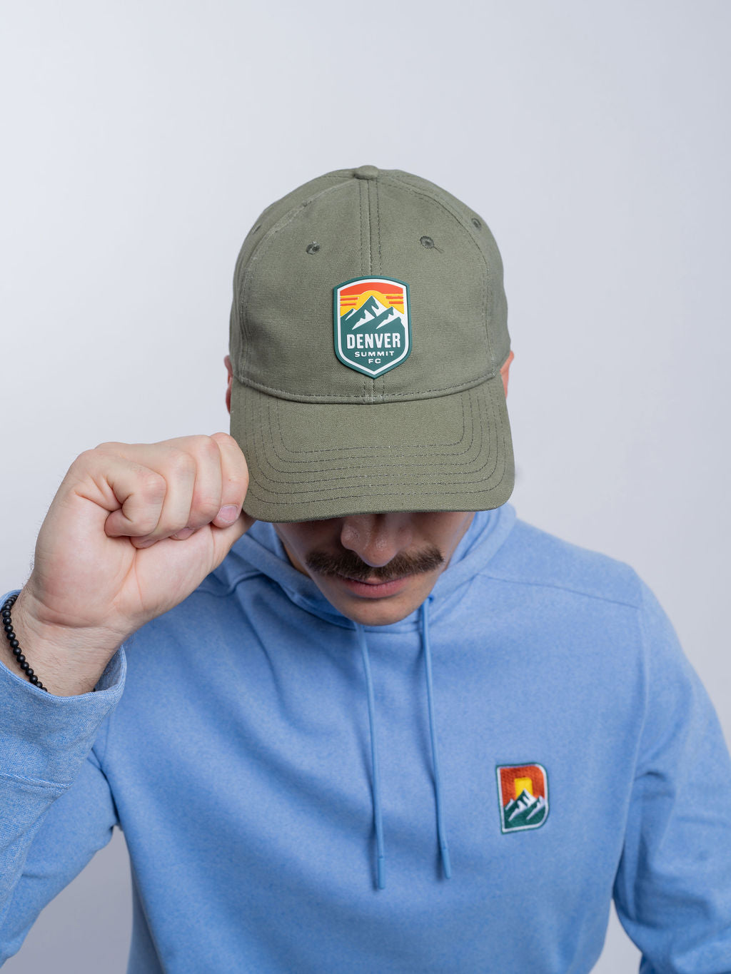 Person wearing a green cap and blue hoodie with logos, on a plain background